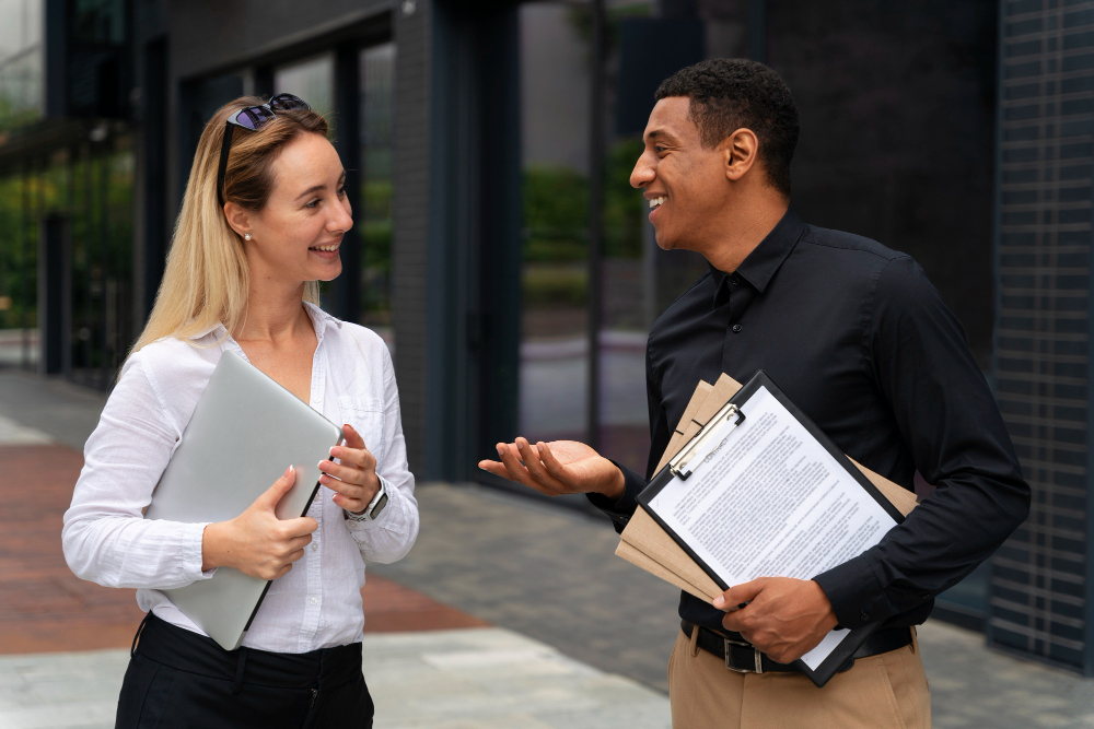 Profissional de recolocação profissional conversando com um candidato ao lado de um prédio, com um laptop e documentos em mãos.