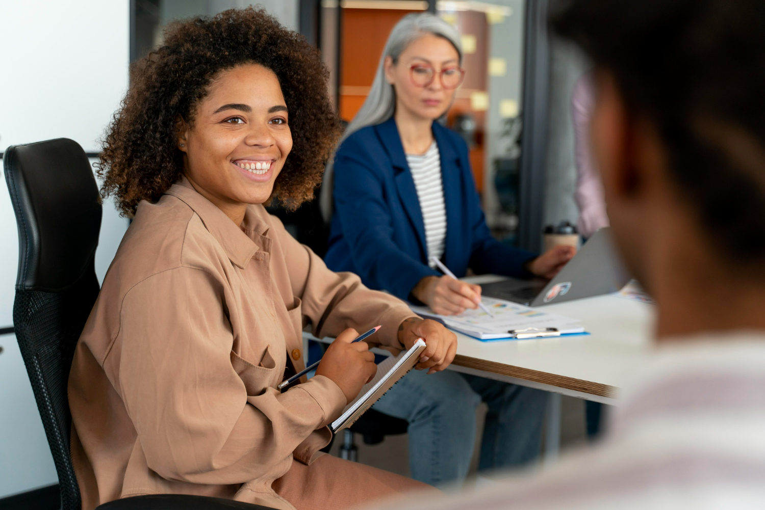 Mulher sorrindo durante reunião de recrutamento para multinacionais, com colegas ao fundo.